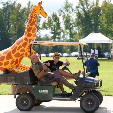 Photo of men on golf card with inflated giraffe. 