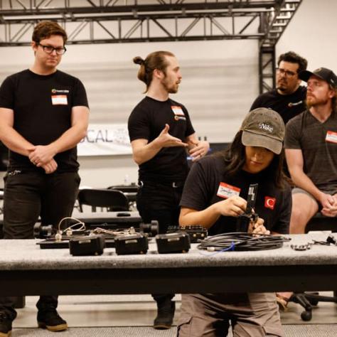 Man sitting at table working with gear as people watch. 