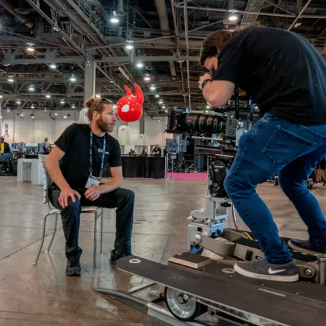 Man hunching over camera, man sitting in front of camera on a chair. 
