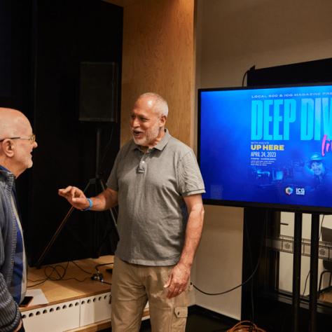 Two men talking in front of a tv screen. 