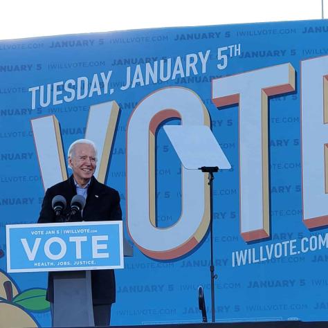 Man speaking at podium in front of 'Vote' sign. 