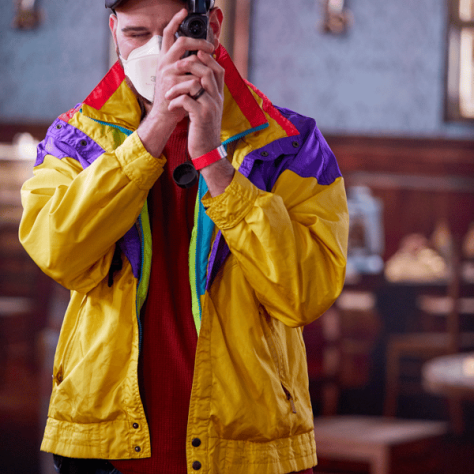 Man with yellow jacket and mask looking into a small camera. 