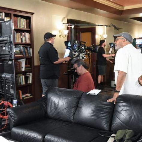 Man standing behind a couch and man on a camera in the background.