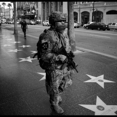 Man in military uniform with gun walking down the street over Hollywood stars. 