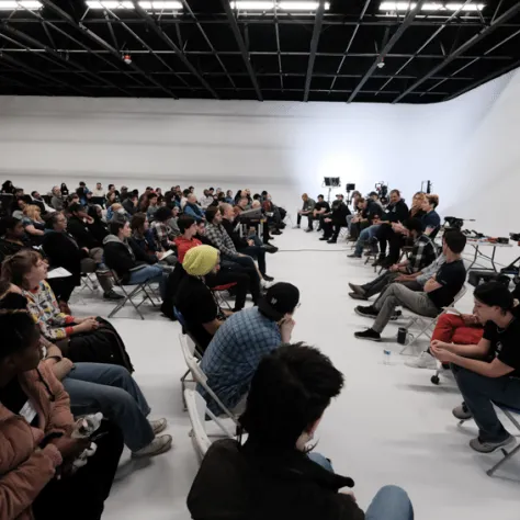 View of audience and people sitting in chairs at the front of the room. 