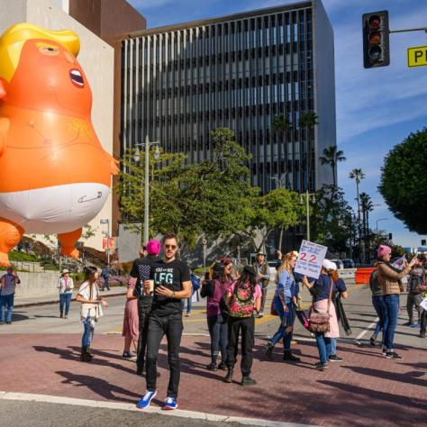 People holding strings attached to a large balloon of a baby Trump in a diaper