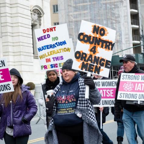 Women marching and holding signs. One reads "DONT NEED NO HATERATION HOLLERATION IN THIS PRESIDENT" and another reads "NO COUNTRY 4 ORANGE MEN" while the rest read "STRONG WOMEN STRONG UNIONS IATSE"