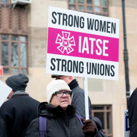 Woman in crowd holding sign that reads "STRONG WOMEN STRONG UNIONS IATSE"
