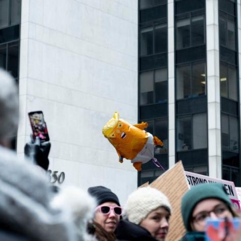 People marching while holding signs and a balloon of a baby Trump in a diaper is floating above