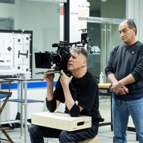 Man sits holding a camera and another behind him watches