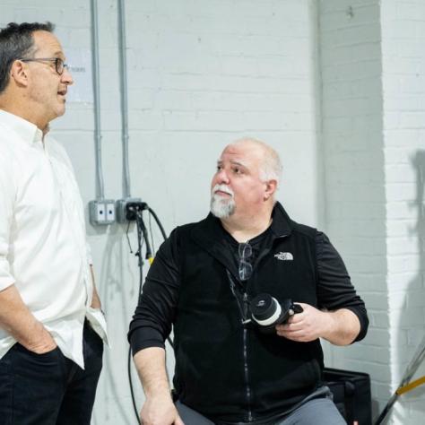Man standing talks to a seated man with a camera