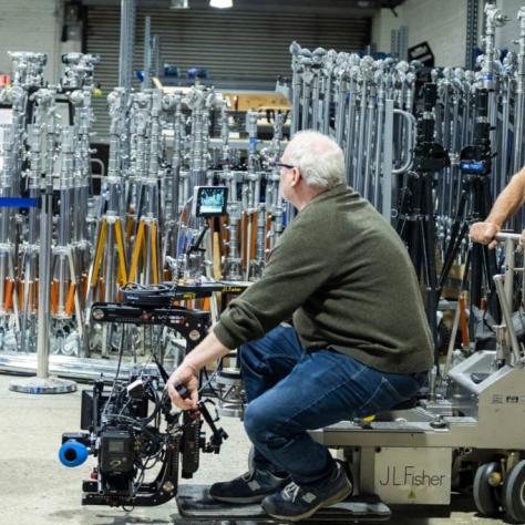 Man sits on wheeled device with camera and another man push it